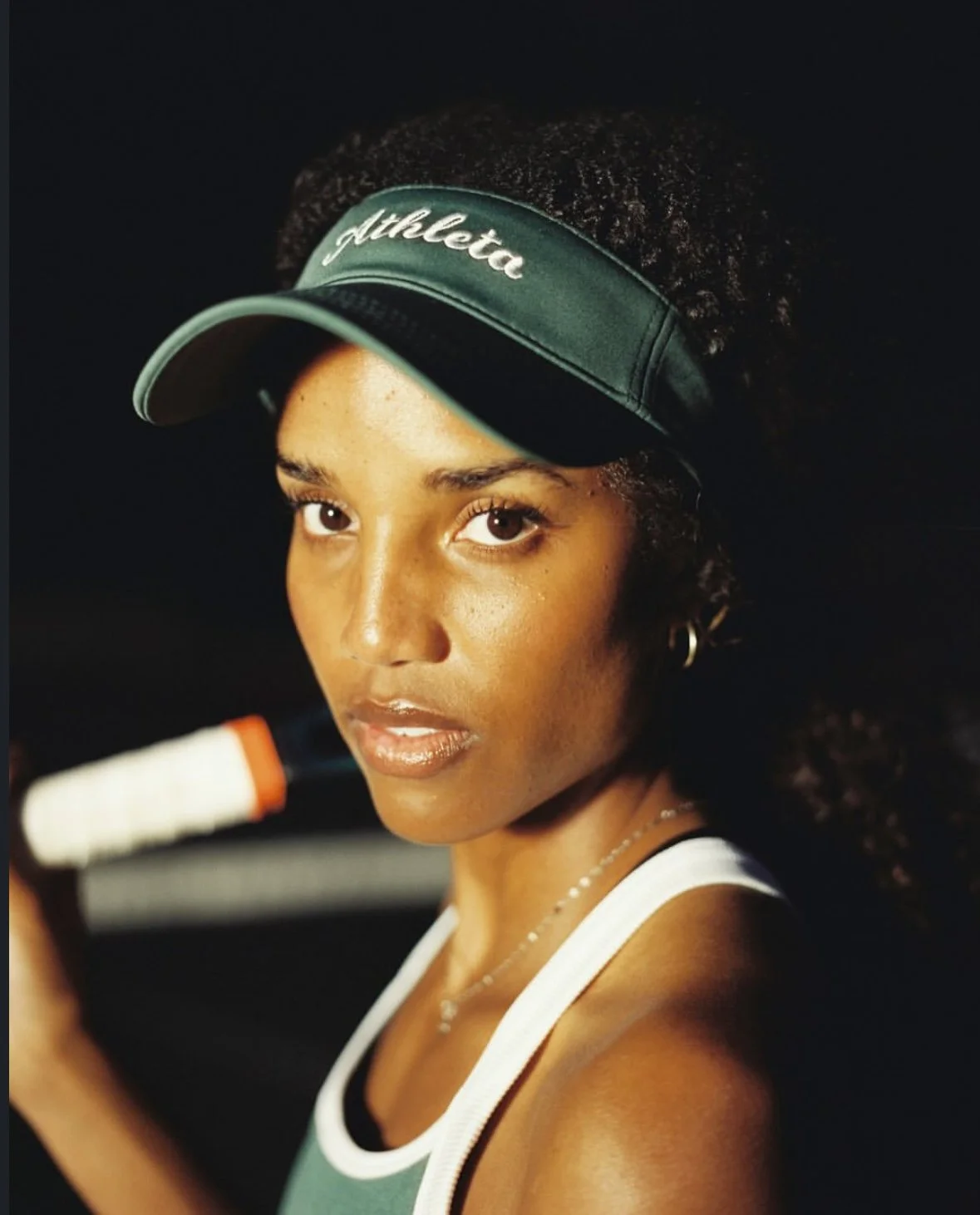 A young woman wearing a green athletic visor and sportswear, holding a tennis racket, looking at the camera with a serious expression.
