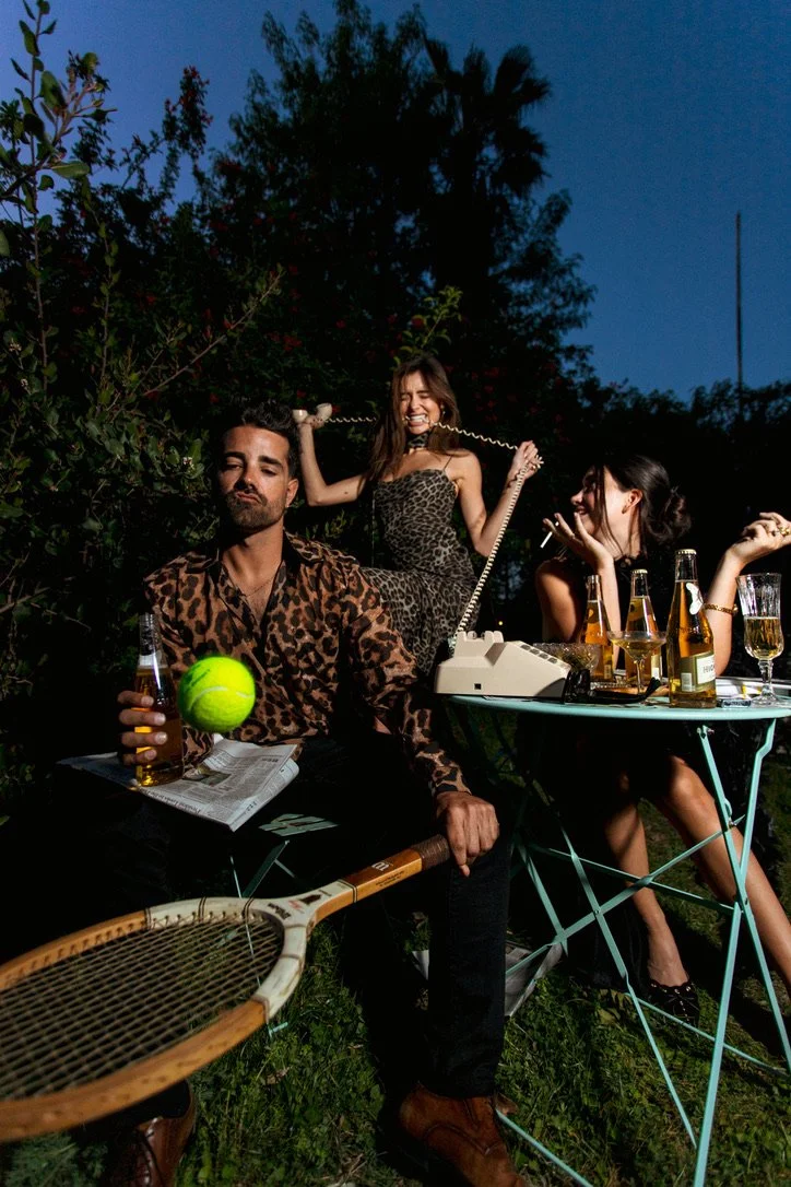 Three young adults having a cocktail party outdoors in the evening with trees in the background, involving tennis, a vintage telephone, and drinks.