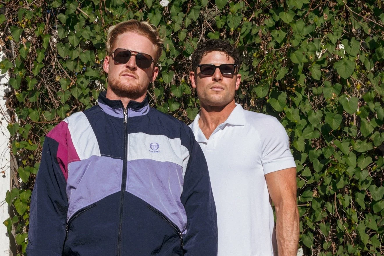 Two young men wearing sunglasses stand outdoors in front of green leafy vines, with one in a vintage color-blocked windbreaker jacket and the other in a white polo shirt.