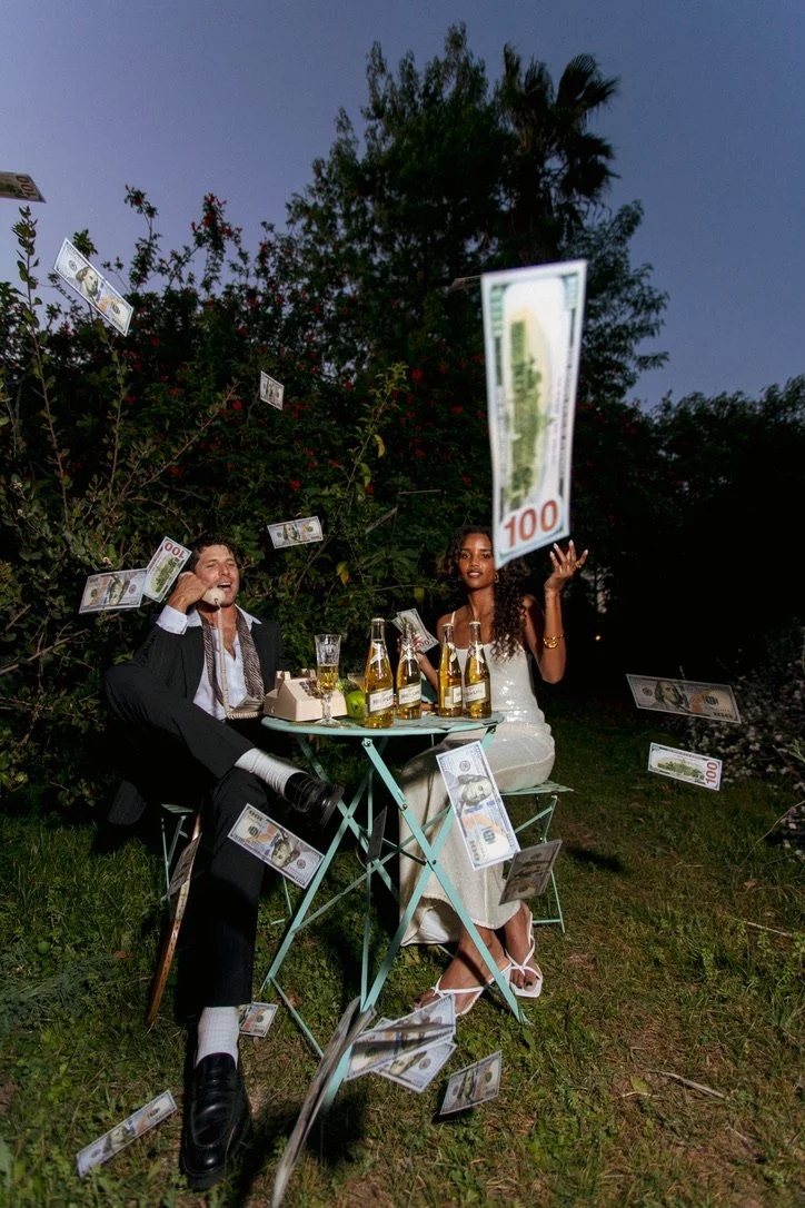 Two people sitting at a small table outdoors, surrounded by flying dollar bills, with empty beer bottles and glasses on the table, under a night sky with trees in the background.