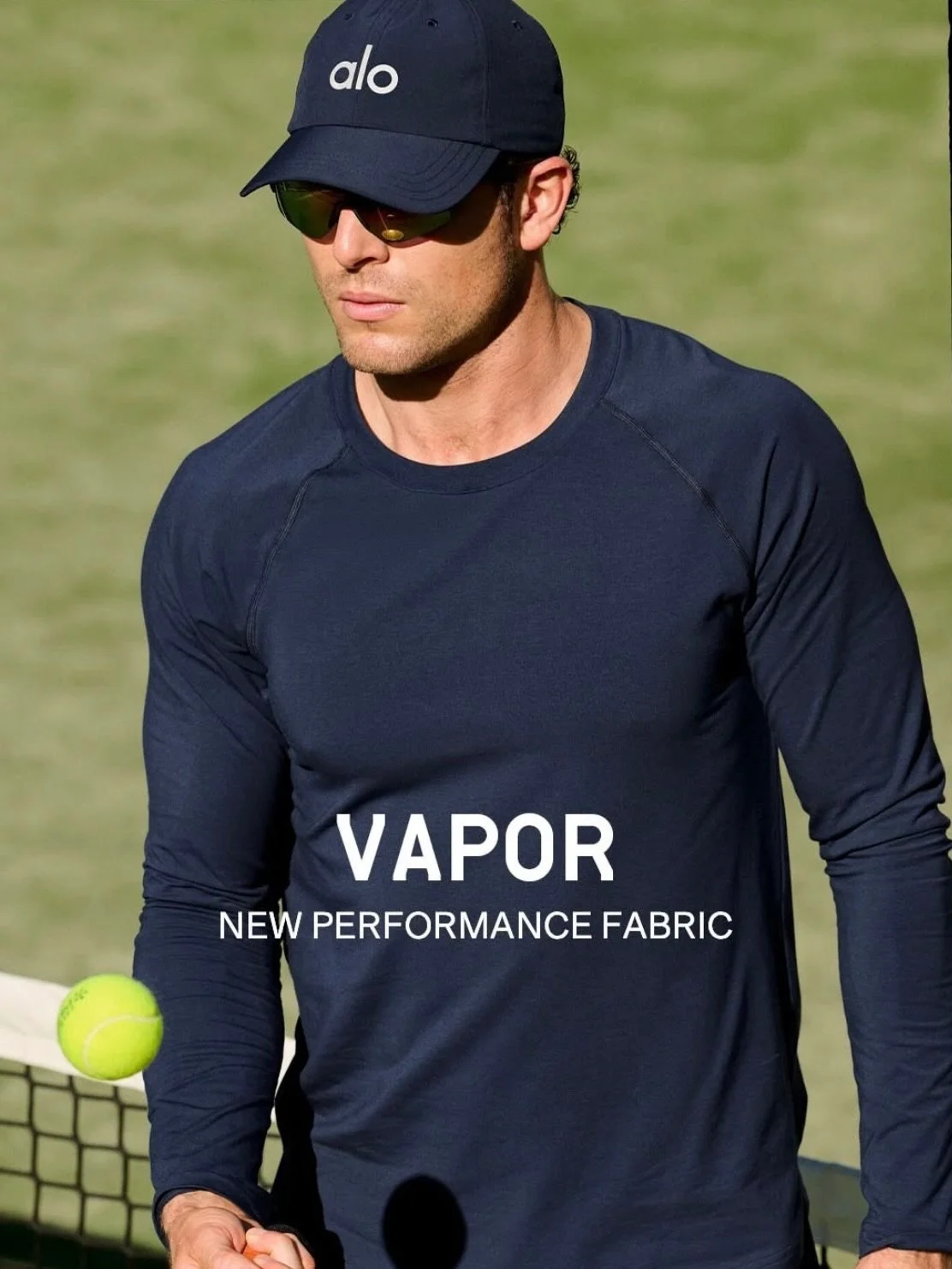 Man wearing a navy blue cap, sunglasses, and a navy blue long-sleeve athletic shirt standing on a tennis court, holding a tennis ball, with a tennis net in the background. Text on the shirt reads "VAPOR NEW PERFORMANCE FABRIC."