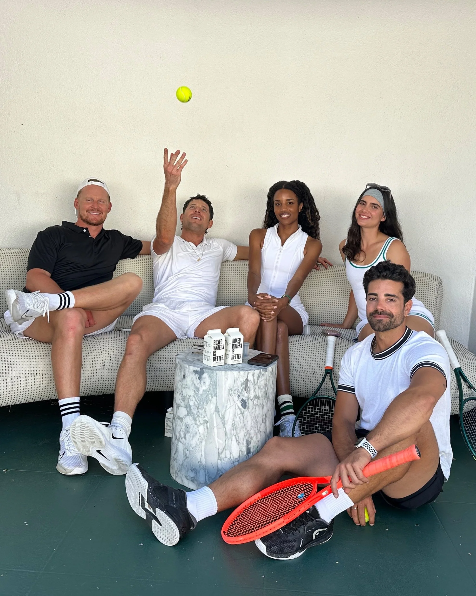 Group of five friends sitting on a sofa and floor, with tennis rackets, a tennis ball, and cartons of water, enjoying a casual moment.