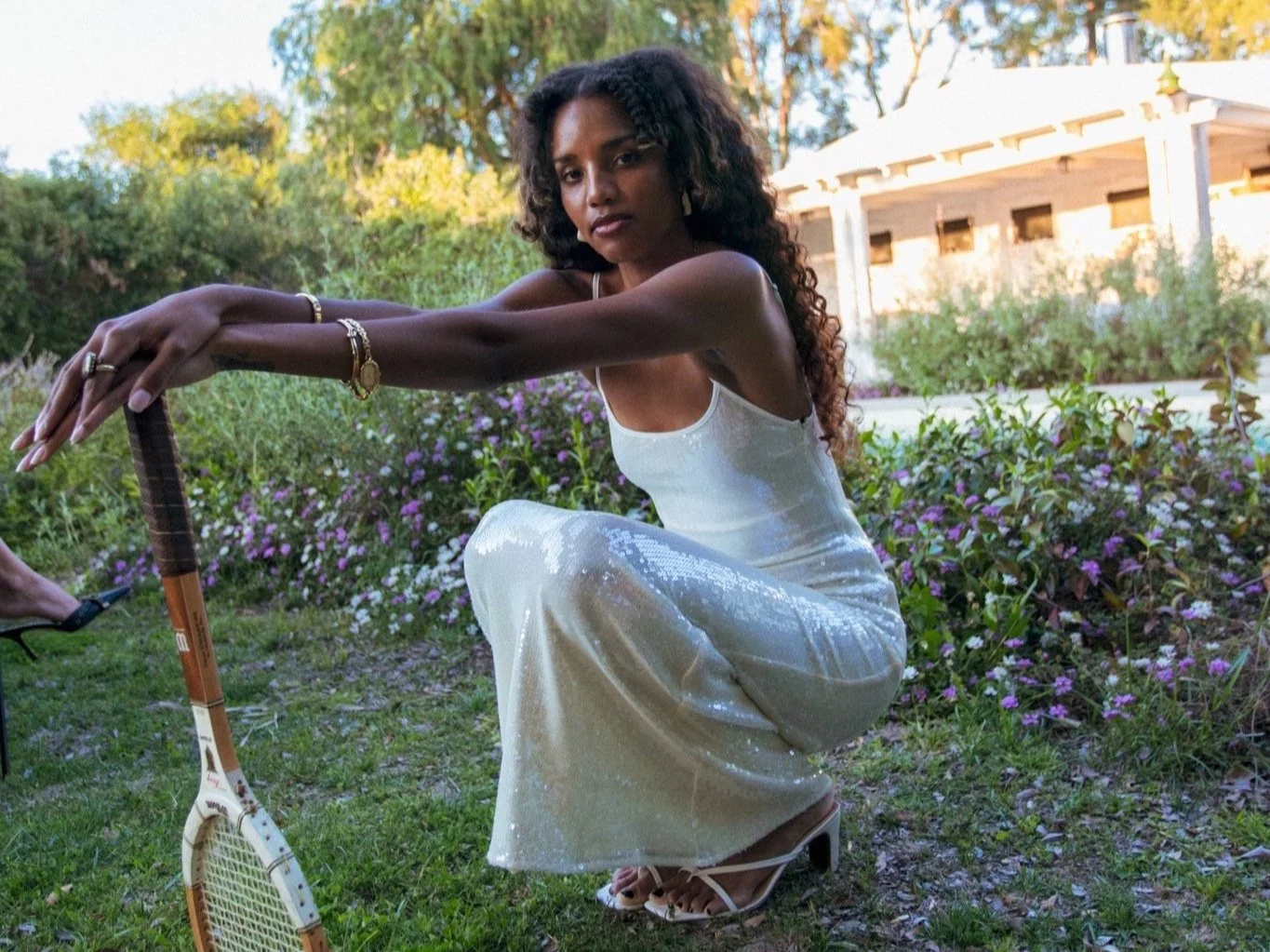 A woman wearing a white, shiny dress, squatting outdoors on grass and purple flowers, holding a tennis racket with her arms extended, with a garden and white house in the background.