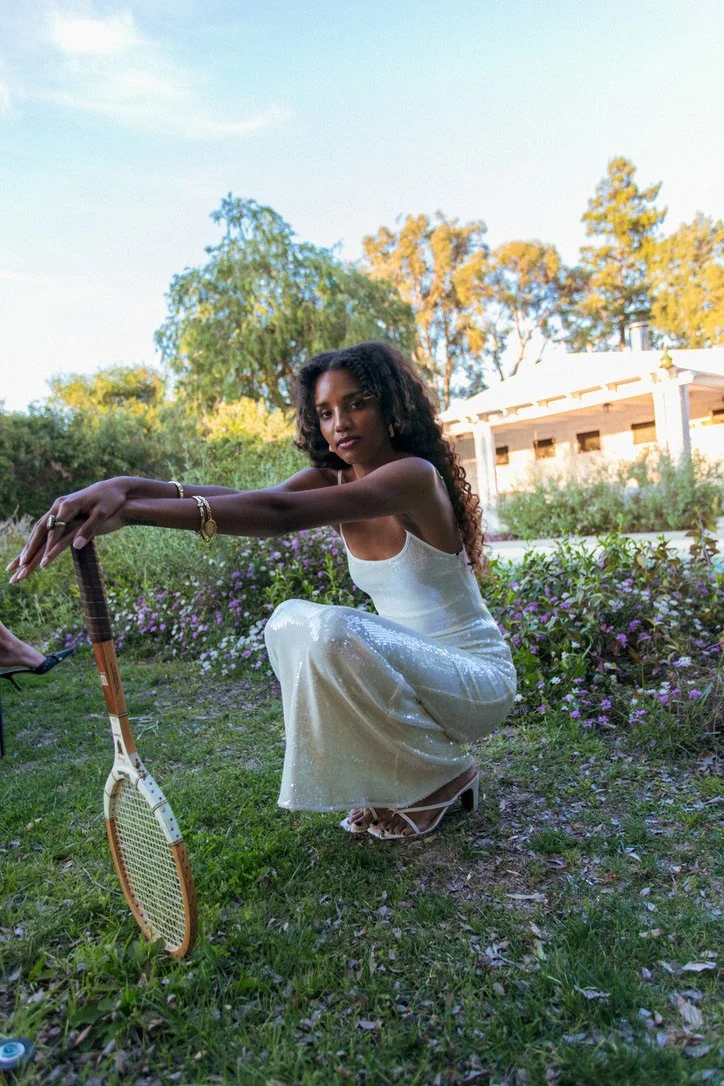 A woman in a white dress crouches on grass, holding a tennis racket in a garden with flowering bushes and trees, with a house in the background.