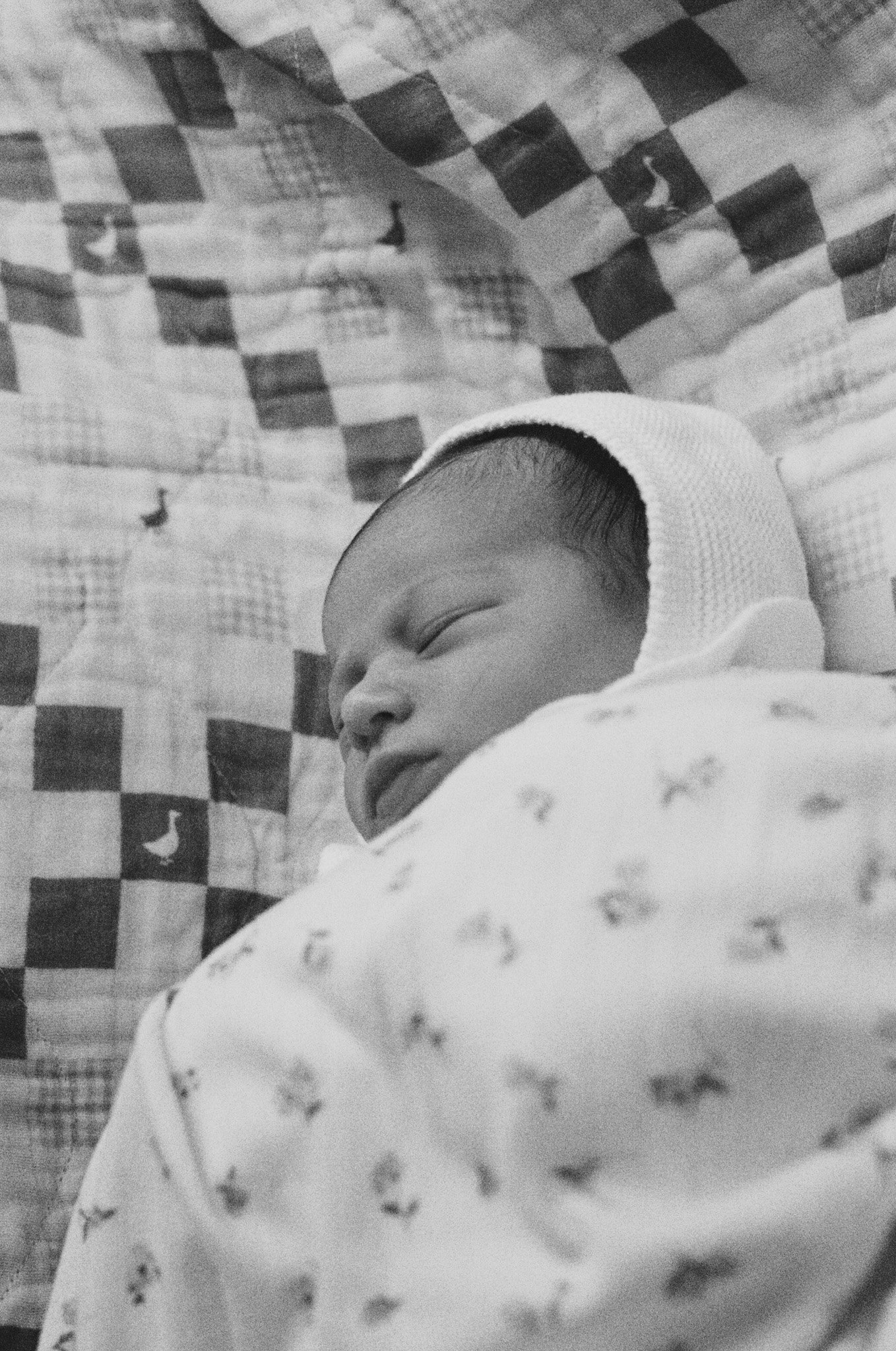 A sleeping newborn baby wearing a white hat and patterned clothes, lying on a checkered blanket.