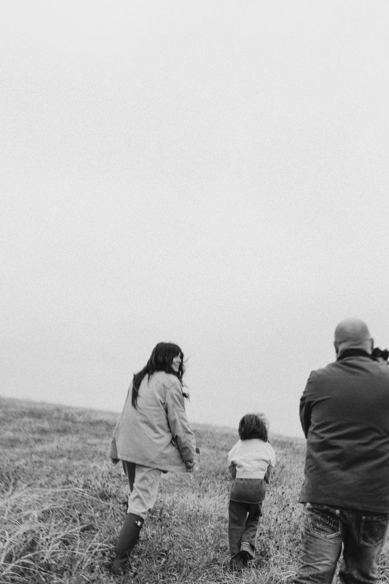 A black and white photo of three people walking through a grassy field, viewed from behind. The group includes a woman, a young child, and a man, with the sky above them.