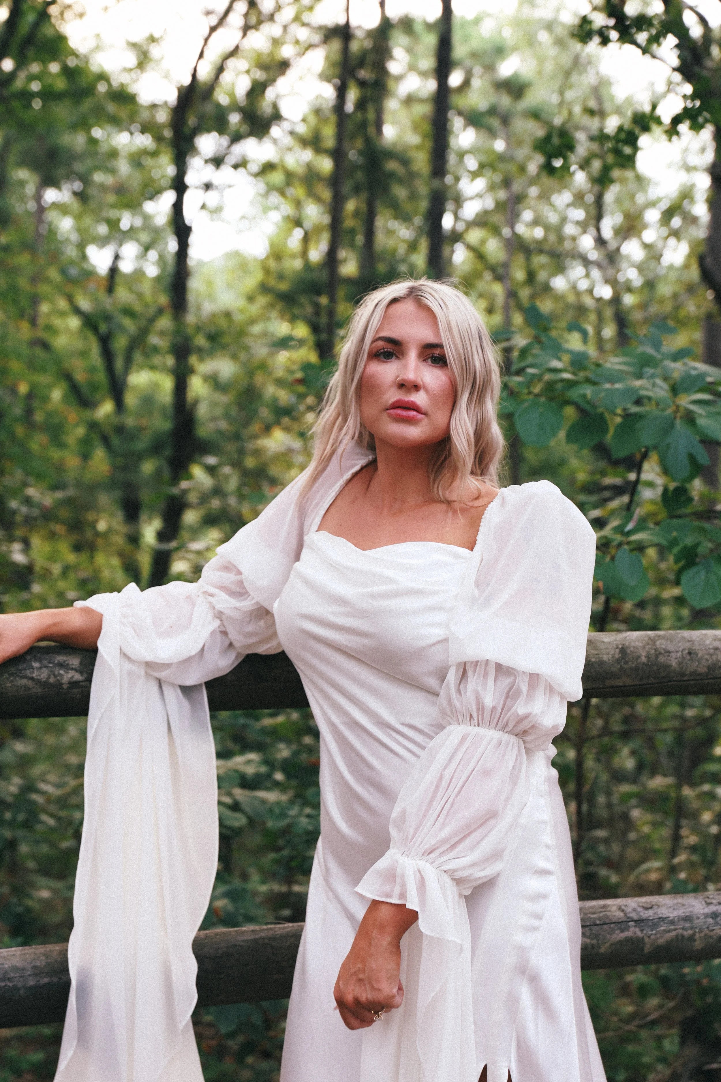 A woman with blonde hair and a serious expression wearing a white dress with puffy sleeves is standing outdoors in a forest with green foliage and trees.