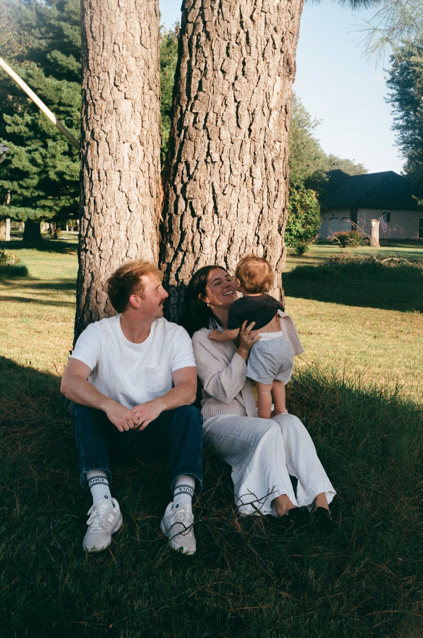Three people, a man, a woman, and a child, sitting and playing under two large trees in a grassy yard, smiling and interacting with each other. on 35mm film