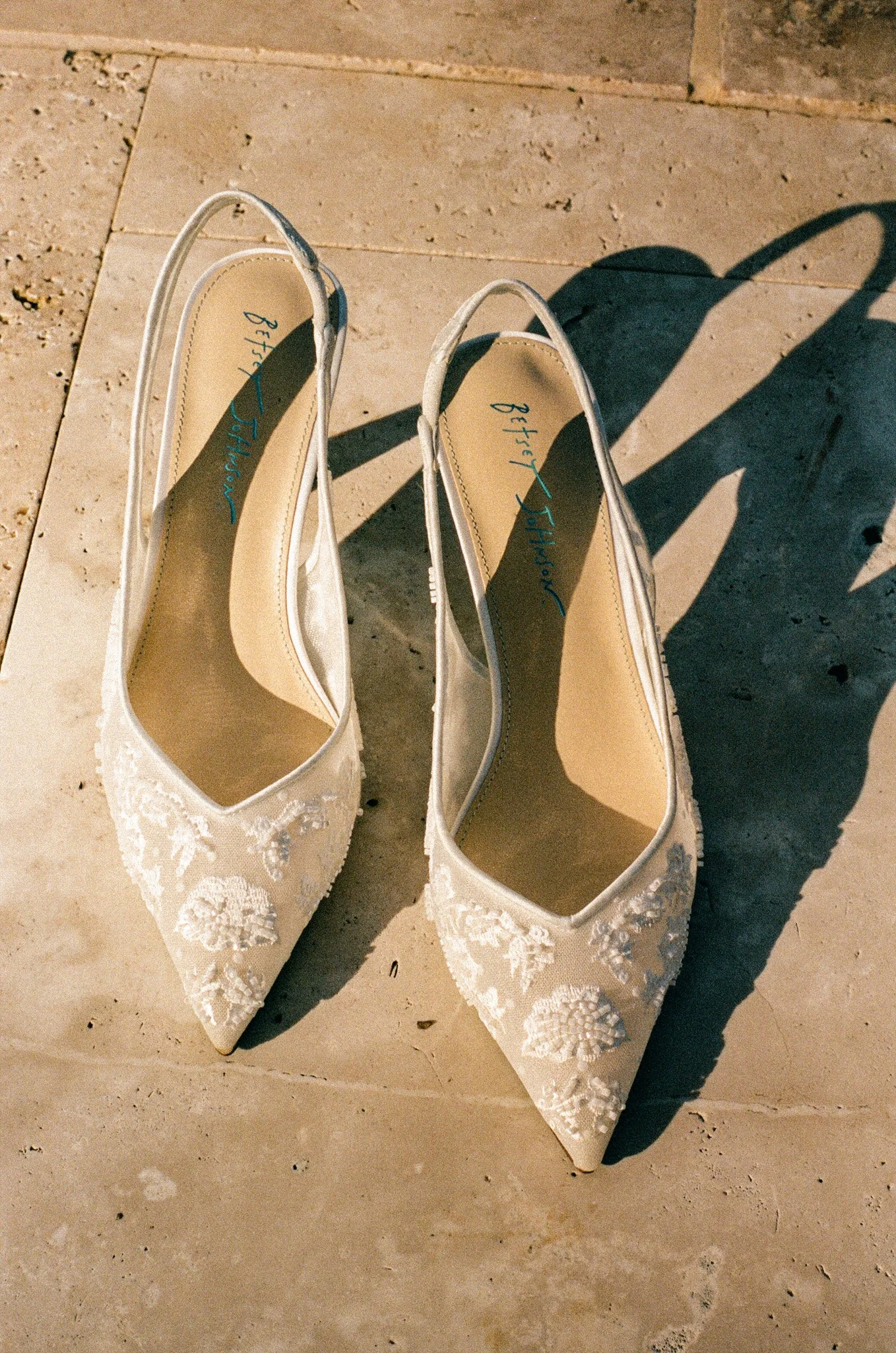 A pair of white bridal shoes with pointed toes and lace embroidery, placed on a tiled floor with sunlight casting shadows. on 35mm film.