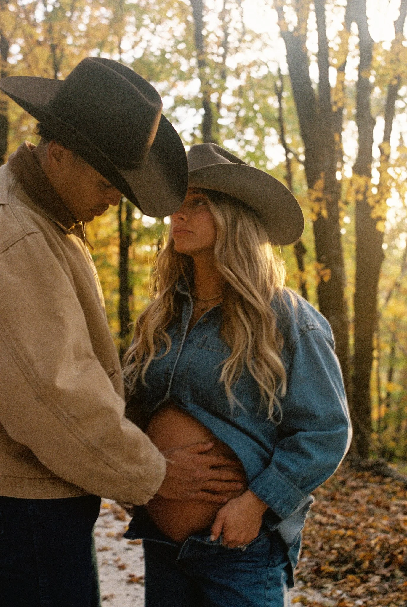 A couple wearing cowboy hats in a wooded area, with the man holding the woman's pregnant belly. on 35mm film.
