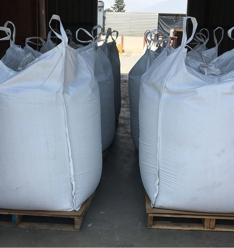 Large white and gray bags on wooden pallets inside a warehouse or storage area, with an outdoor scene visible in the background.