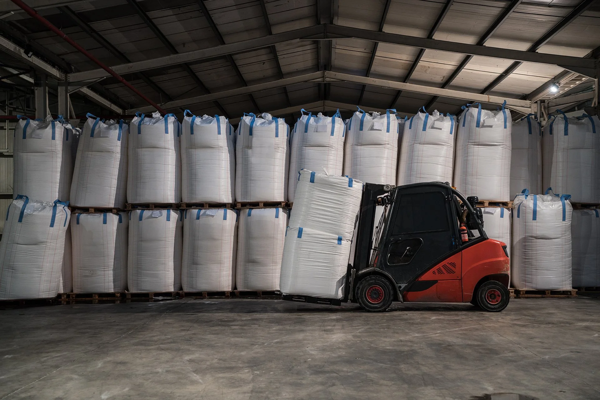 A red forklift lifting a large white sack in a warehouse with stacked similar sacks in the background.