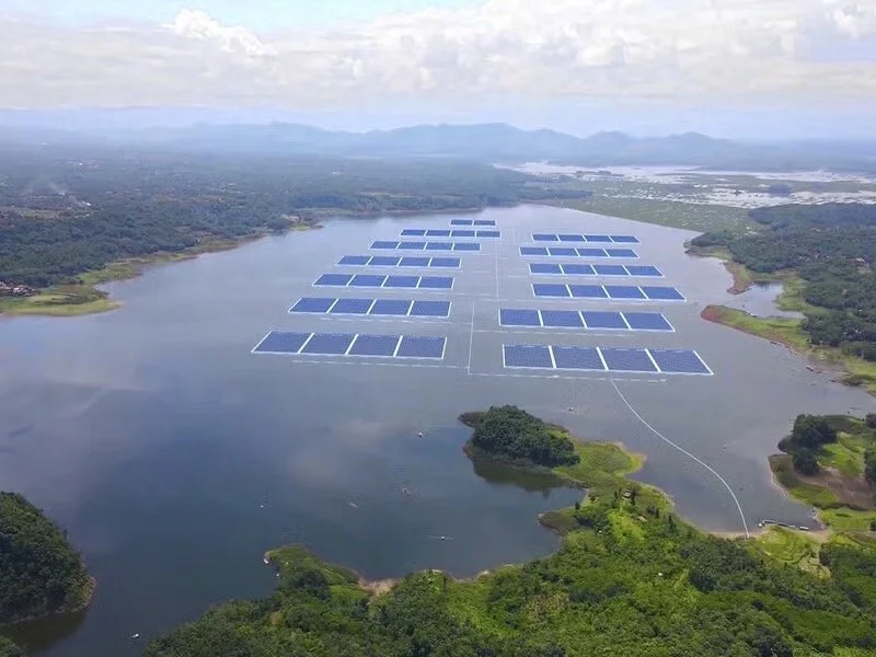 Aerial view of a lake with multiple floating solar panels creating a solar farm, surrounded by green landscape and distant mountains in the background.