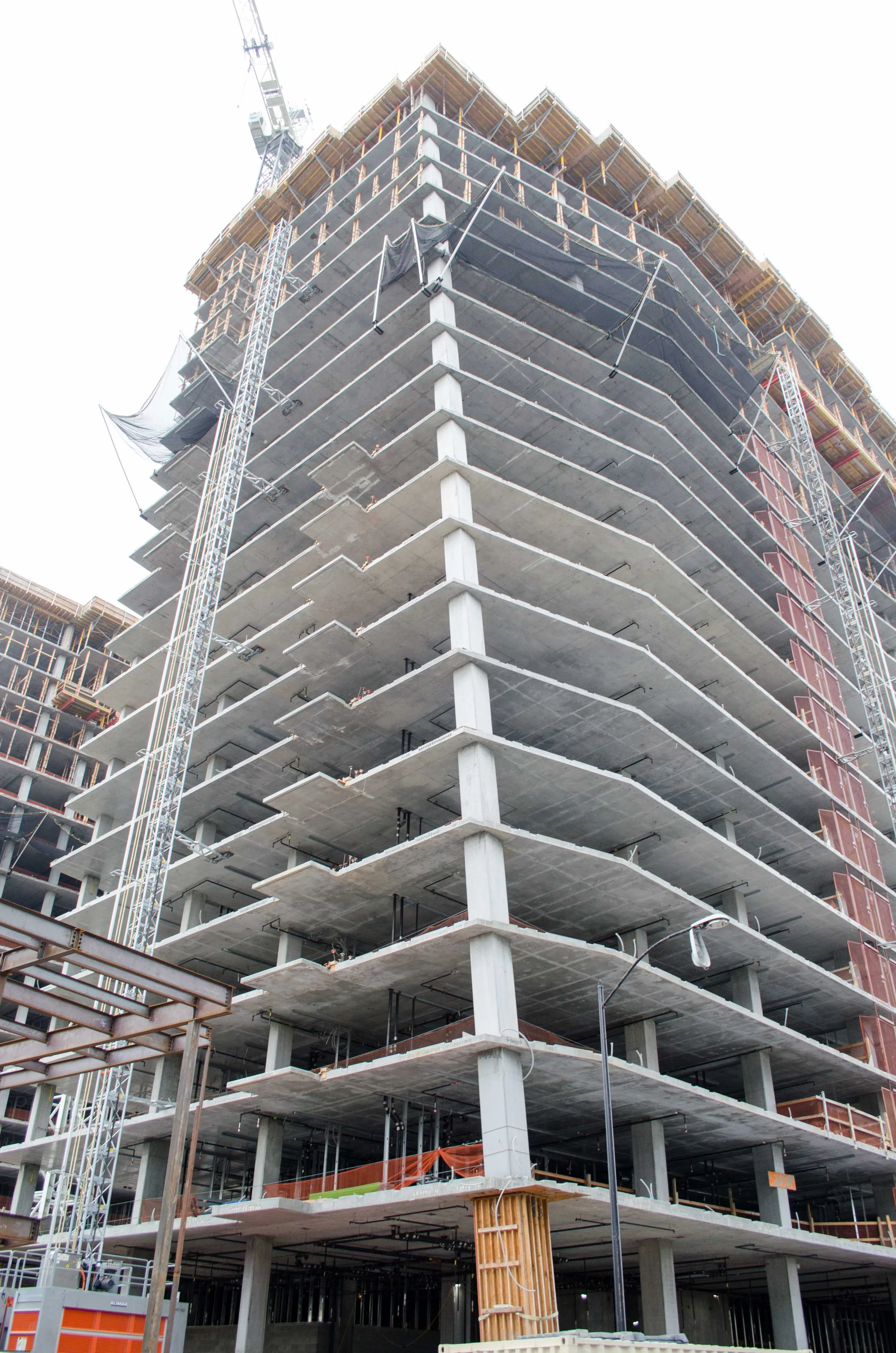 A high-rise building under construction with concrete floors and steel supports, surrounded by scaffolding and construction equipment.