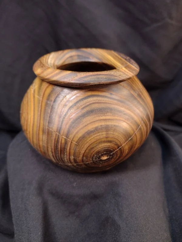 A small wooden vessel with a round base and a wide mouth, featuring natural wood grain patterns in various shades of brown.