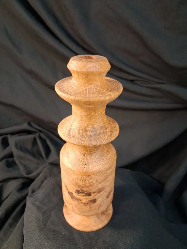 A wooden sculpture with a cylindrical base and three rounded, bulbous shapes stacked on top, carved from a single piece of wood, against a dark fabric background.