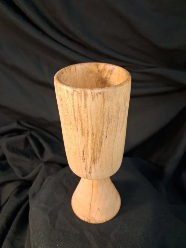 A wooden chalice with a light natural finish, featuring a smooth, rounded cup and a wider tapered base, set against a black fabric background.