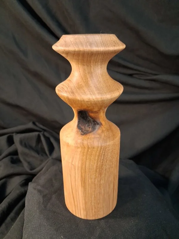 A carved wooden sculpture with a smooth, natural finish, featuring a rounded base, an hourglass shape, and a narrow top, set against a black fabric background.
