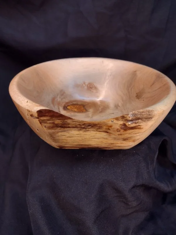 A wooden bowl with a natural, uneven finish and visible wood grain, placed on a black fabric background.