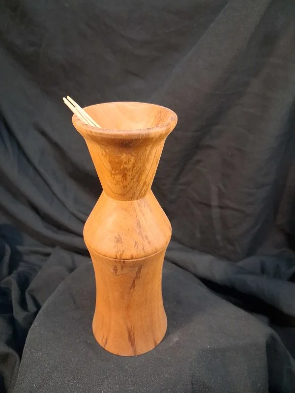 A wooden vase with an hourglass shape, holding two pairs of chopsticks, placed on a black fabric background.