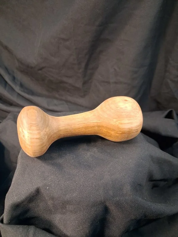 A wooden massage hammer with rounded ends resting on black fabric against a dark background.