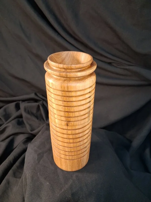 A tall, cylindrical wooden object with evenly spaced horizontal grooves, set against a black fabric background.
