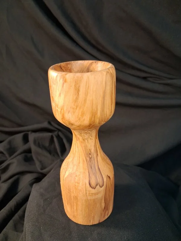 A wooden cup with a rounded base and a wider opening, made of light-colored, smooth, polished wood, placed against a dark fabric background.