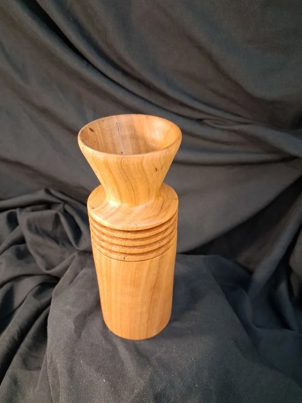 A wooden vase with a curved, flared opening and a smooth, cylindrical body, placed against a dark fabric background.