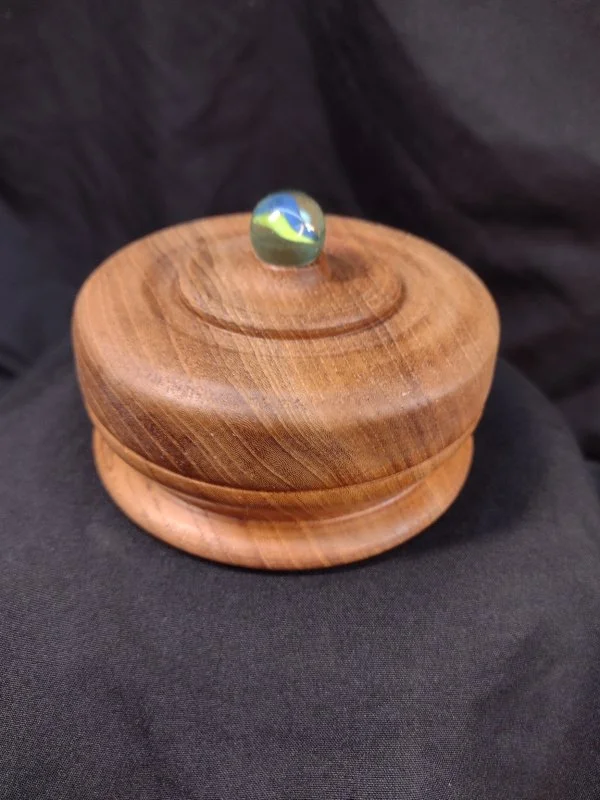 Wooden container with a round lid and a glass knob on top, placed on black fabric.