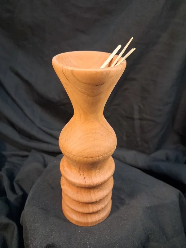 A wooden sculpture resembling a tall vase with a bulbous middle section, holding a few light-colored chopsticks against a dark fabric background.