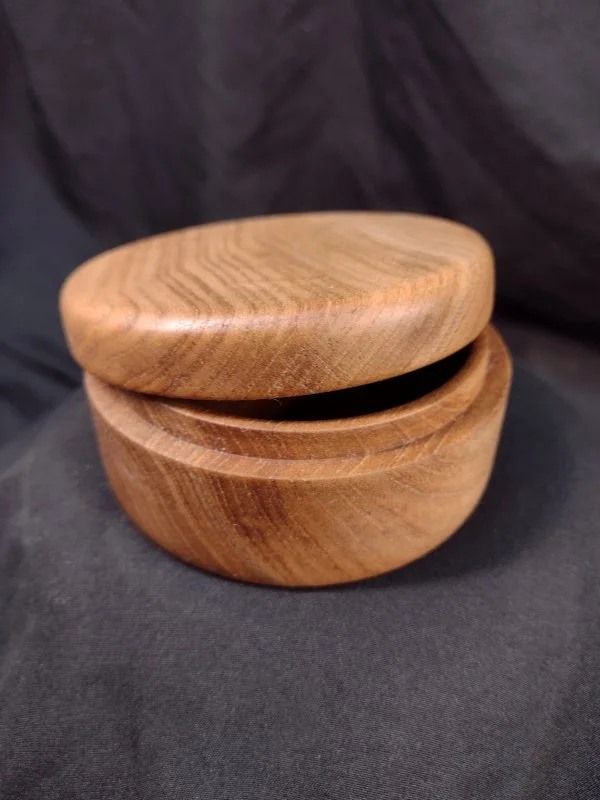 Round wooden container with a lid, possibly a storage box or trinket box, placed on a black fabric background.