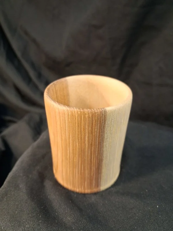 A wooden cylindrical cup with vertical grain patterns, sitting on a dark fabric surface.