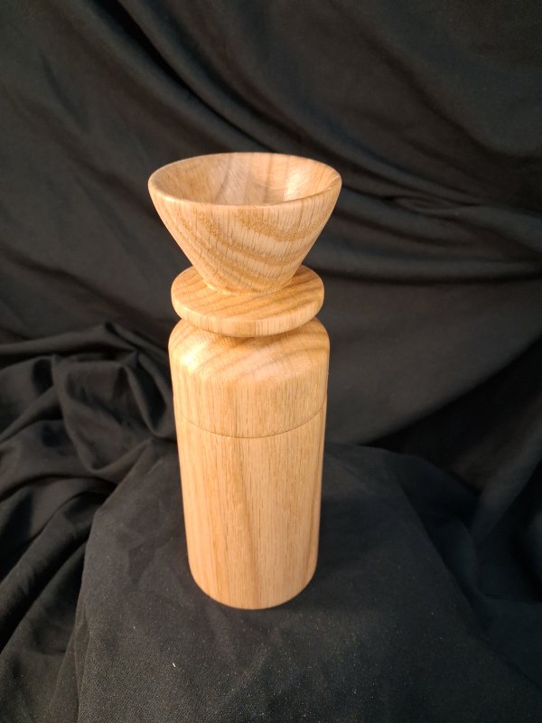 A wooden pepper grinder with a rounded top, set against a dark fabric background.