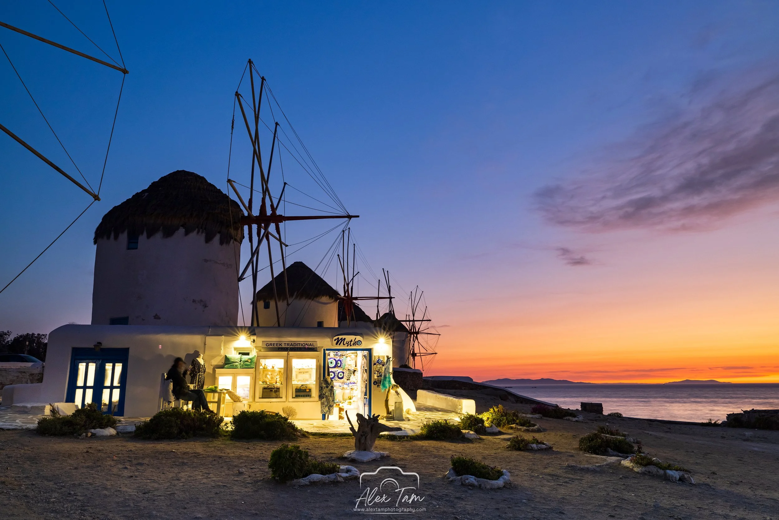 Traditional white windmill with thatched roof, small shop with illuminated sign, and a person sitting outside at sunset overlooking the ocean.