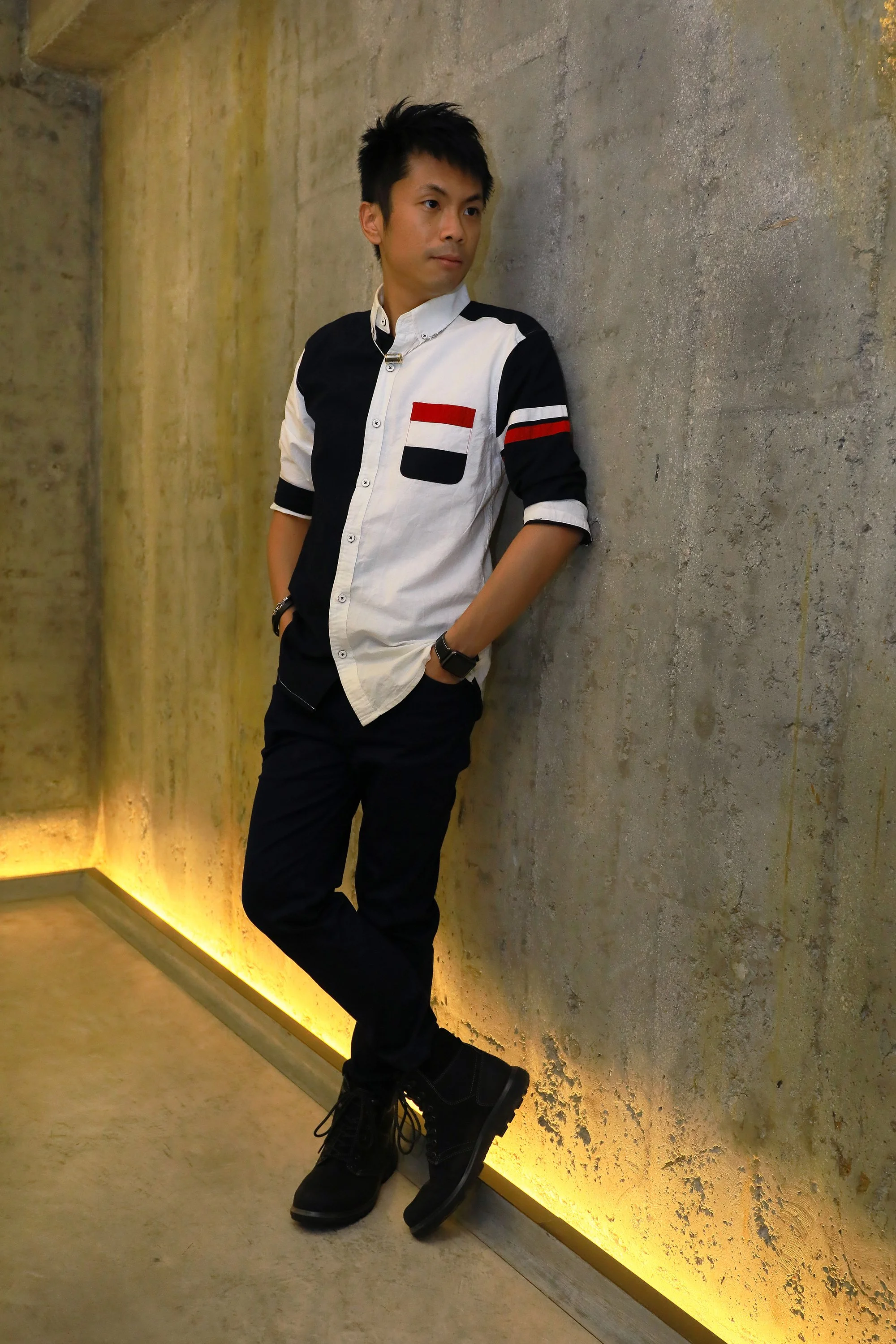 Young man leaning against a concrete wall with hands in pockets, wearing a black and white shirt with red and black accents, black pants, and black boots.