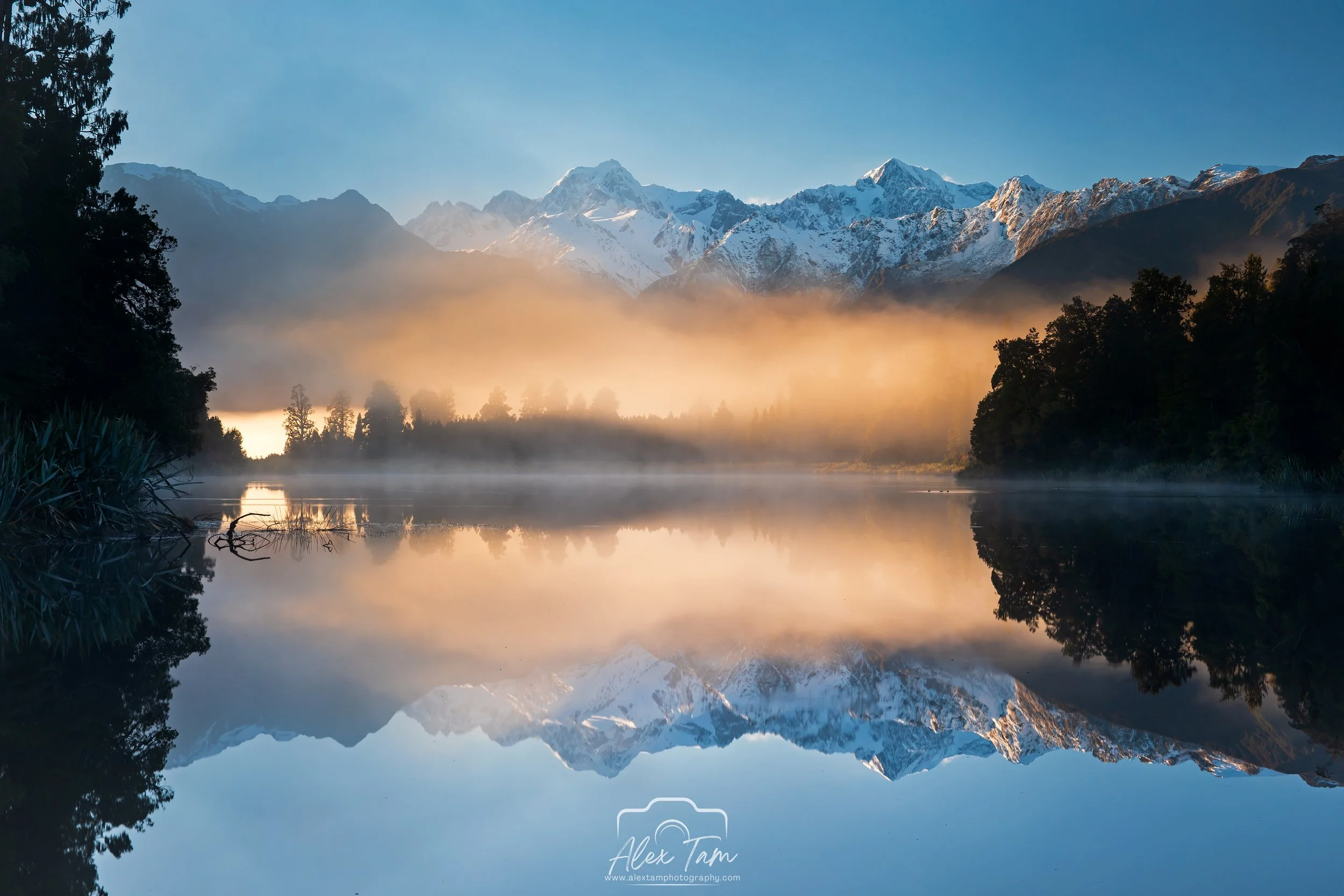 A serene mountain landscape with snow-capped peaks, mist over a calm lake, trees on both sides, and a clear blue sky.