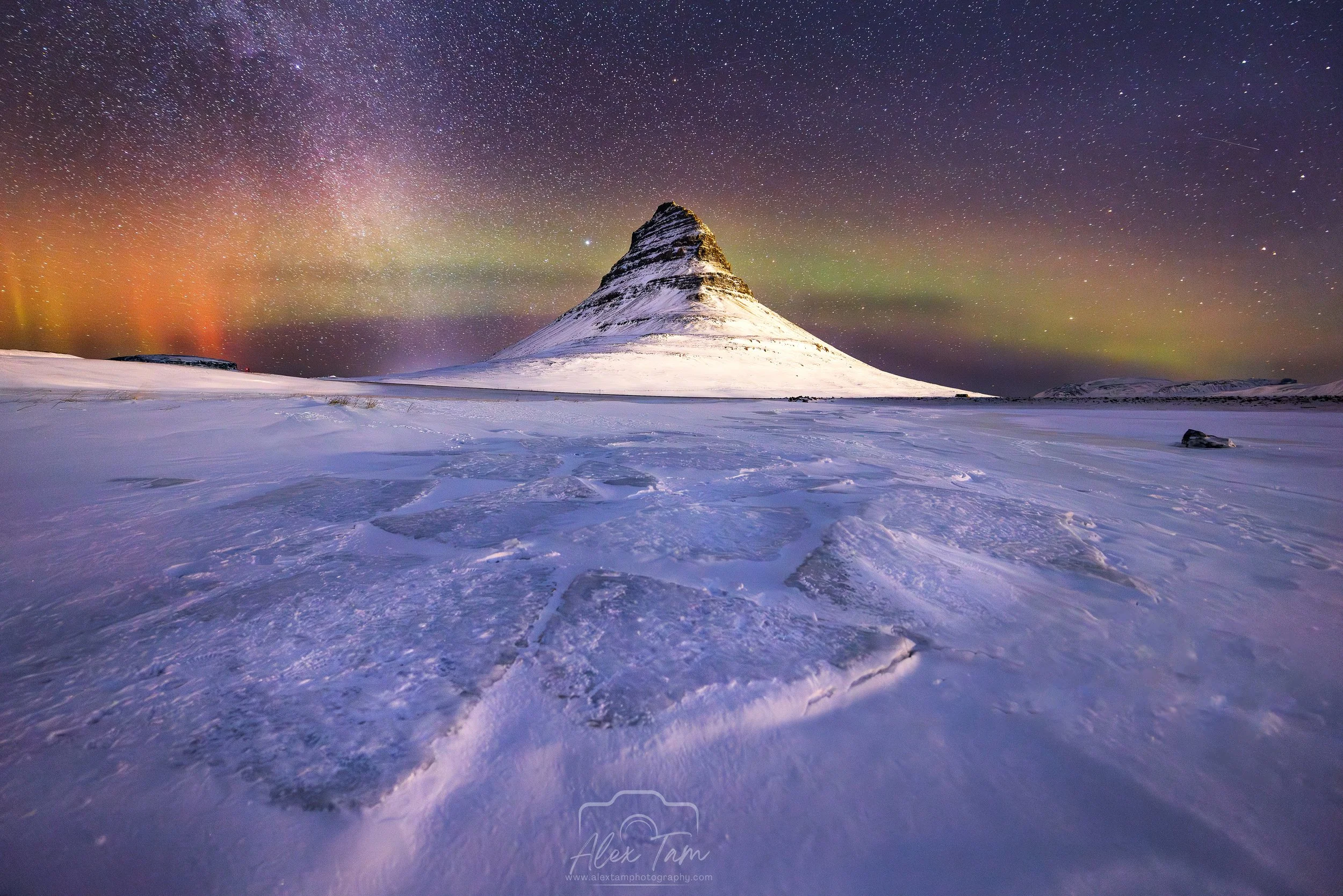 Snow-covered mountain under a starry sky with the northern lights, in a flat icy landscape.