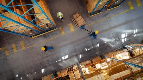Warehouse with workers in safety helmets and vests, walking and handling pallets.