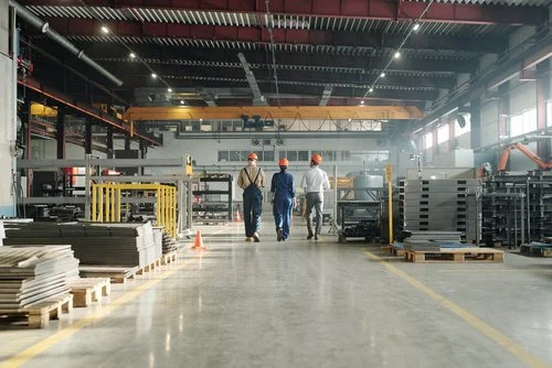 Three workers wearing hard hats walking through an industrial manufacturing warehouse with equipment and supplies on either side.