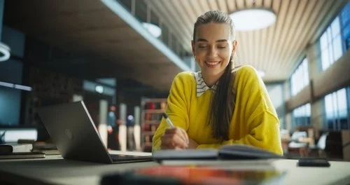 A smiling young woman in a yellow sweater sitting at a desk with a laptop, writing in a notebook in a modern indoor space with large windows.