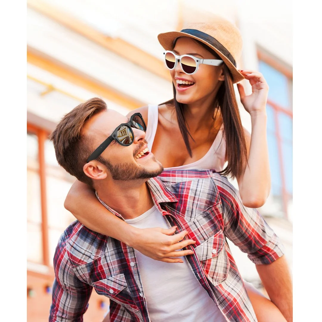 A young man giving a piggyback ride to a young woman, both wearing sunglasses, outdoors with a building in the background.