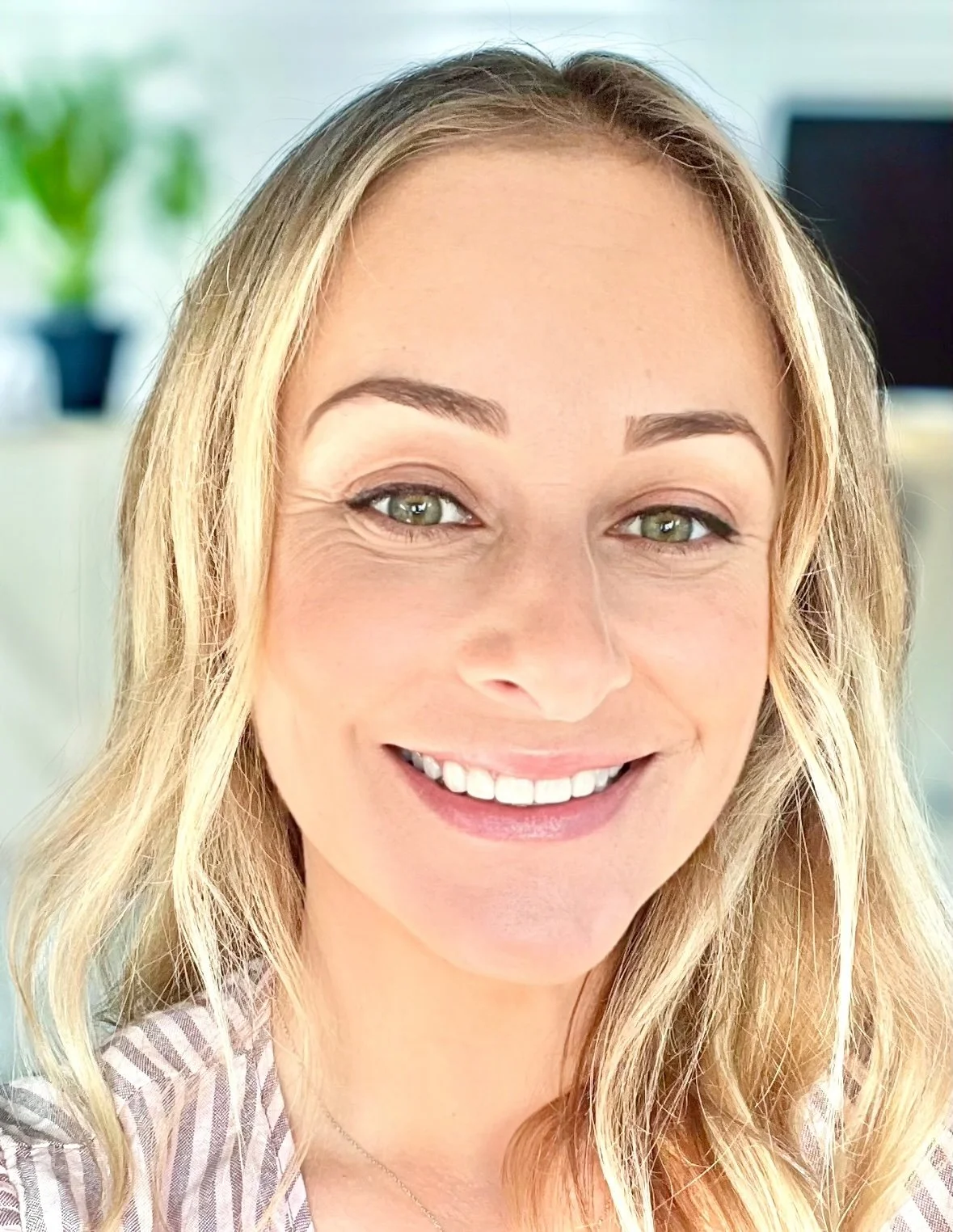 A smiling woman with blonde hair and green eyes, wearing a striped shirt, in a bright indoor setting.