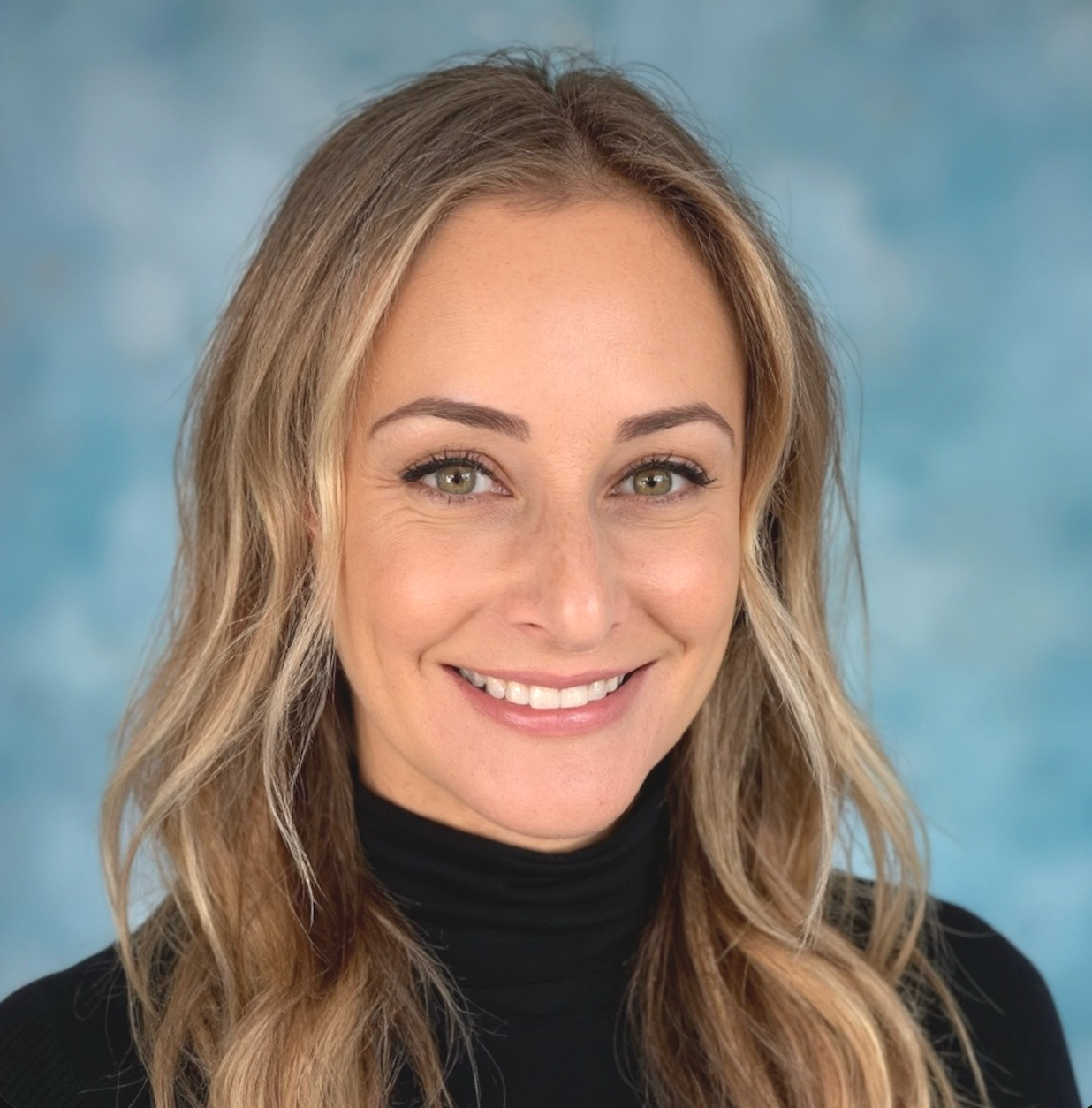 Close-up portrait of a smiling woman with blonde hair and green eyes, wearing a black turtleneck, against a blue sky background.