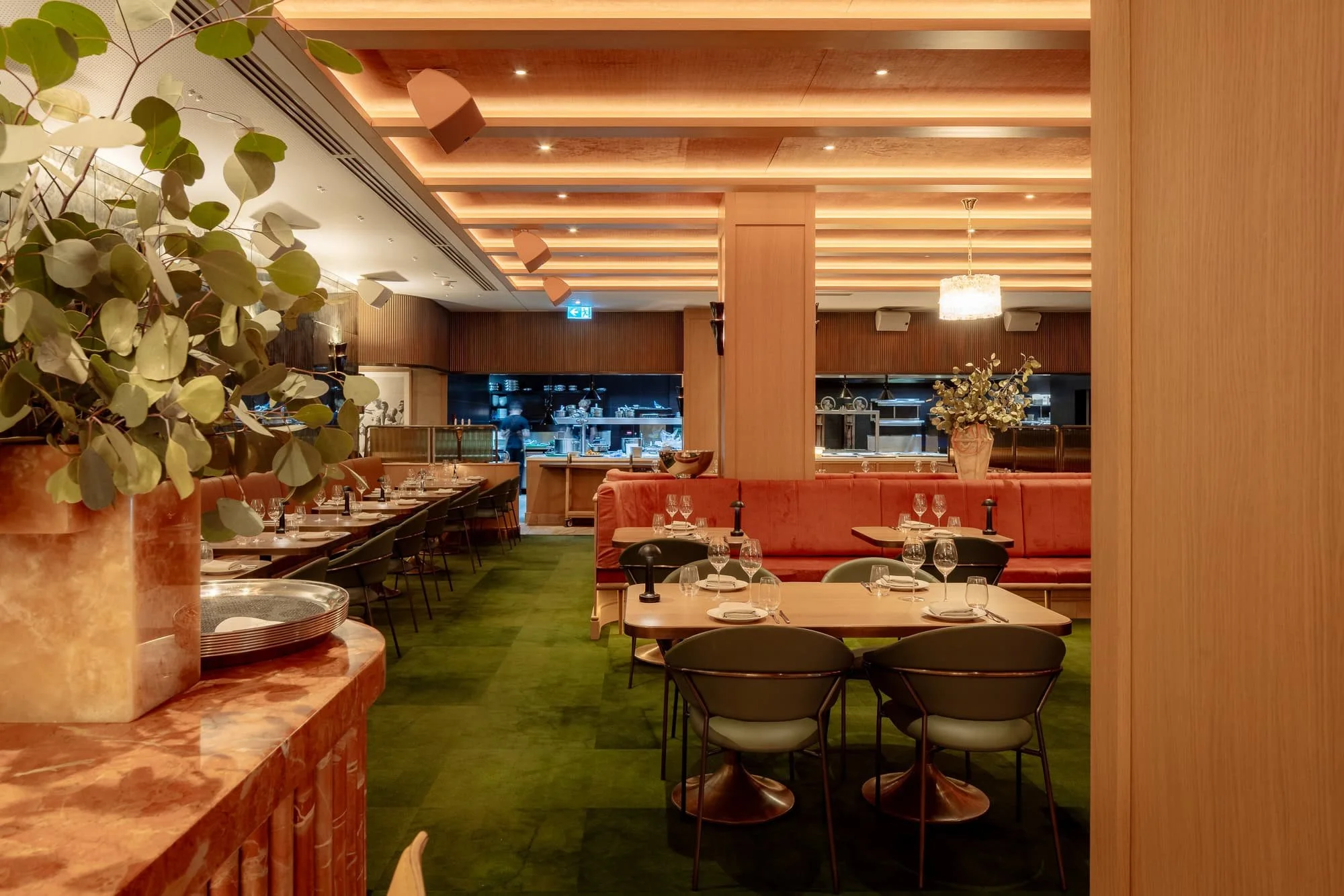 Interior of a restaurant with wooden paneling, green carpet flooring, tables set with glasses and plates, and decorative plants.