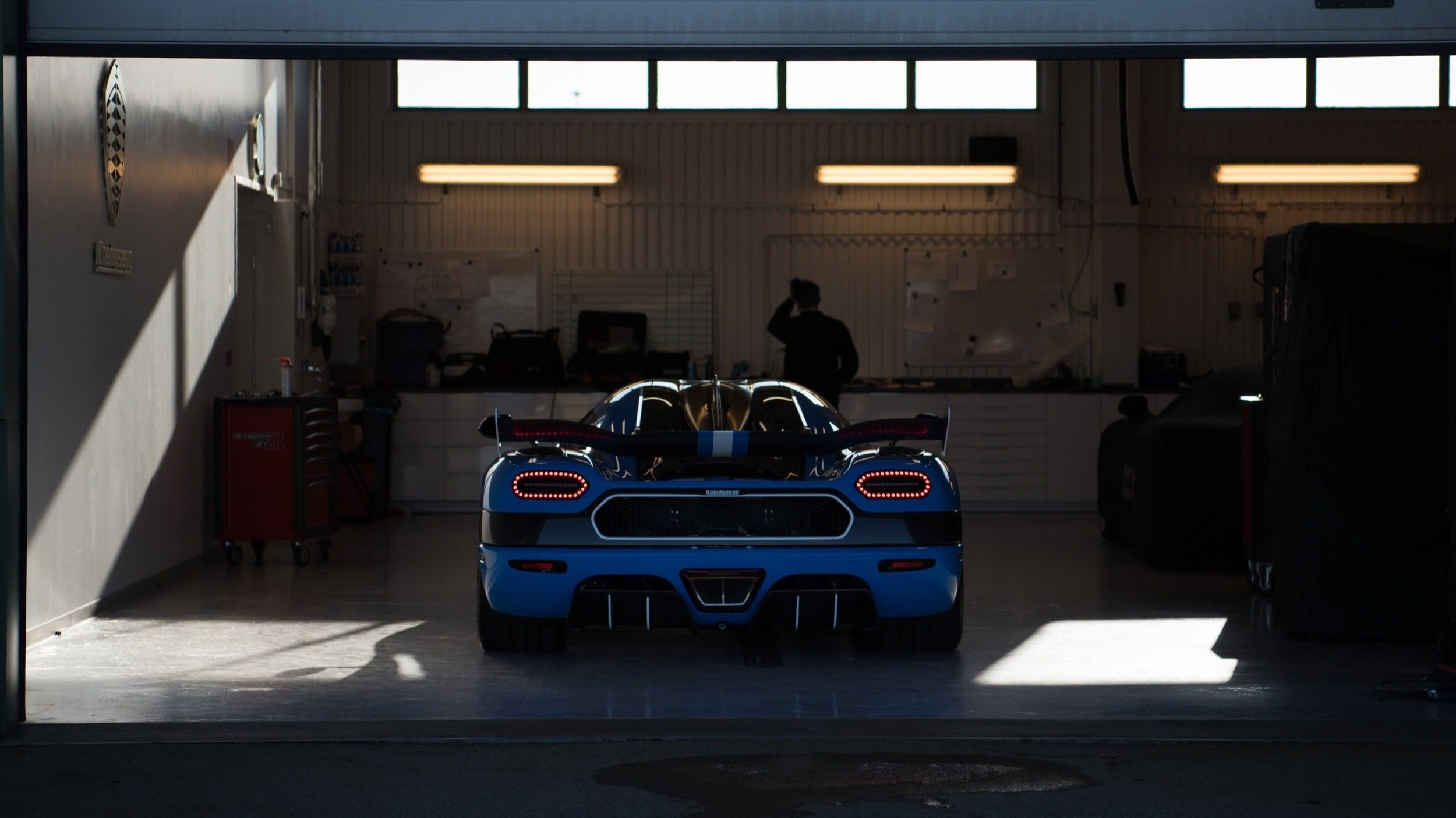 A blue race car inside a garage, viewed from the back, with tools and a person working in the background.