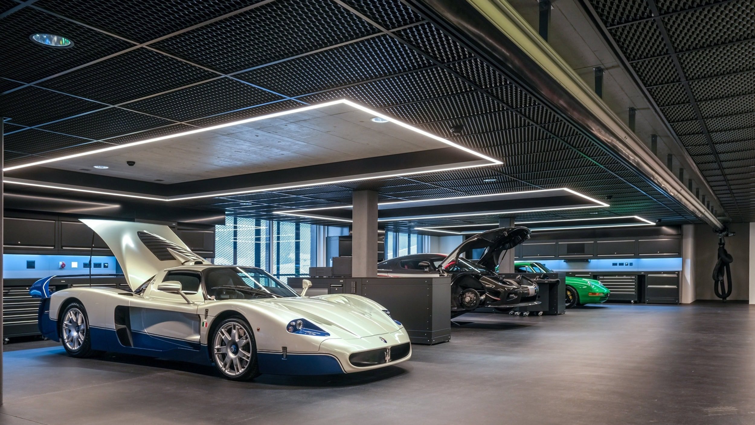 Luxurious car garage with three exotic cars: a silver and blue race car, a black sports car, and a green sports car, under modern ceiling lighting.