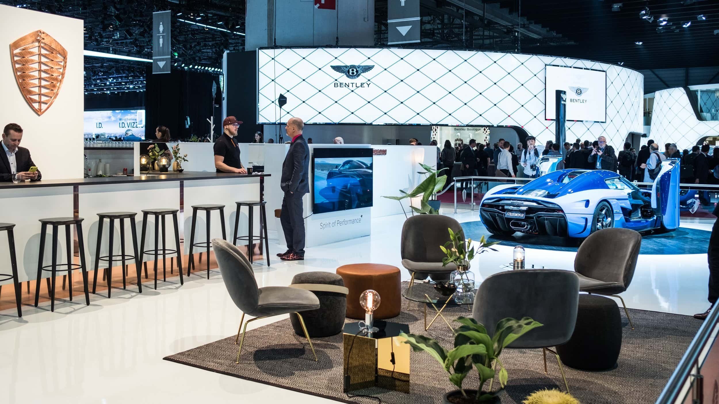 A luxury car display at an auto show with a brightly colored sports car, seating area with modern chairs and small tables, and a bar area with stools. In the background, a large "Bentley" logo is visible on a white geometric backdrop.
