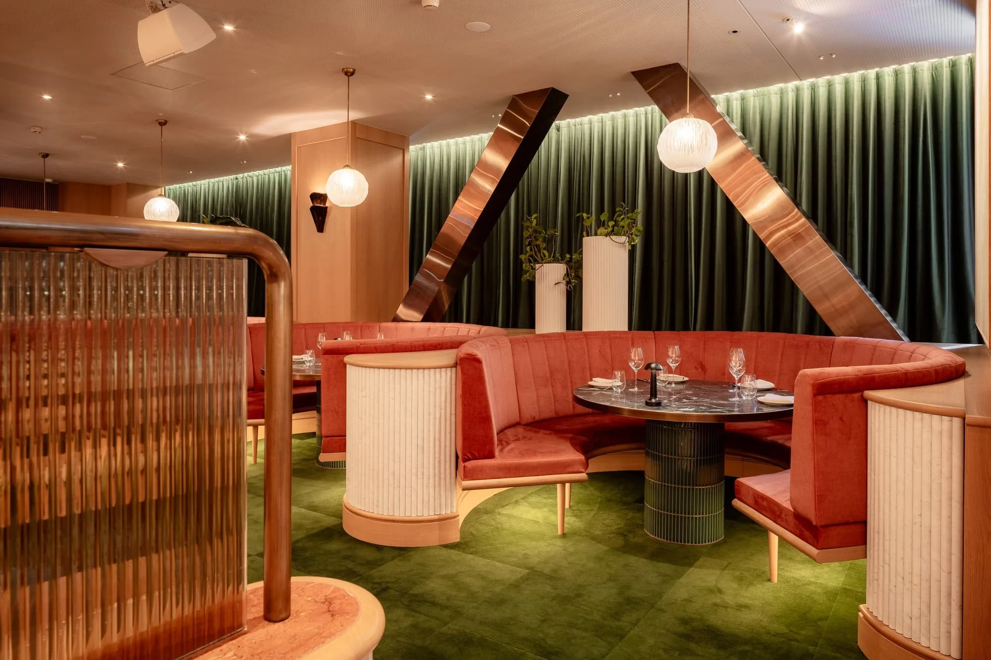 A modern restaurant interior with pink booths, a round black marble table set with wine glasses, tall white planters with green plants, green curtains, and decorative lighting fixtures including pendant lights and wall sconces.