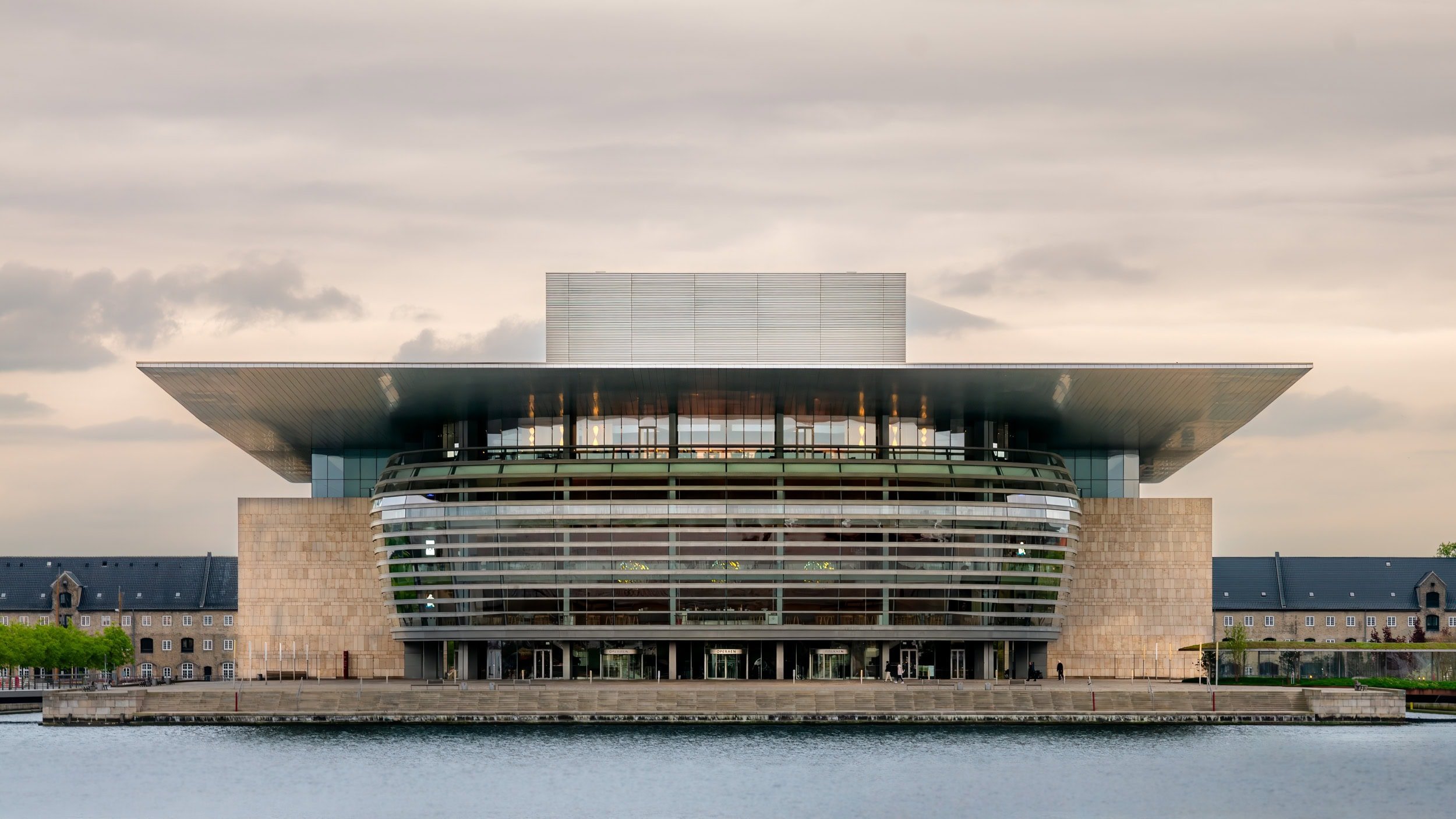 Modern cultural building with curved glass facade, stone walls, large overhanging roof, situated by water, with cloudy sky overhead.