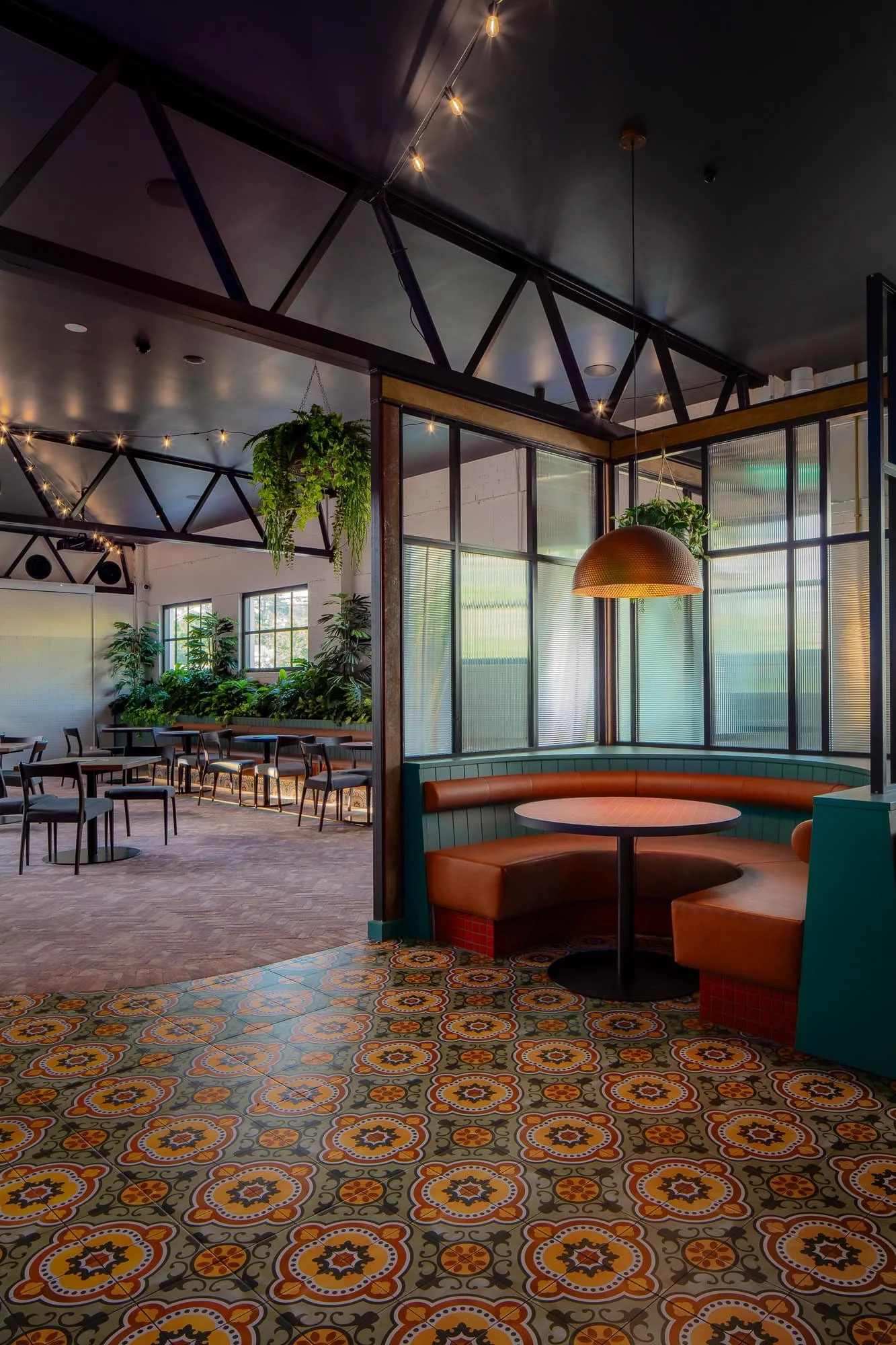 Interior view of a modern restaurant or café with vintage patterned tile flooring, mid-century style seating, abundant green plants, and large windows with natural light.